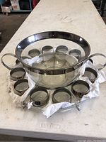 Glass punch bowl with metal rim and handles surrounded by 10 matching cups on a cloth inside their metal holder.