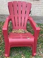 Red/burgundy plastic Adirondack chair shown outdoors on grass, with fallen leaves on seat.