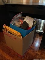 Medium cardboard box filled with assorted office and packing supplies, showing contents visible from the top and sides including padded envelopes, file folders, pens, tape, glue bottles and small boxes.