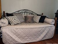 Front view of beige quilted daybed with black metal scrolled back frame, wooden posts, and multiple cushions including square and round bolsters.