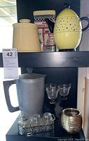 Two shelves holding decorative items including a yellow perforated ceramic teapot, beige ceramic jar, gray pitcher, clear goblets, metallic candle holder and a tray with three small jars