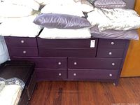 Front view of purple wooden chest with eight drawers and decorative glass-look drawer pulls, top covered with bedding.