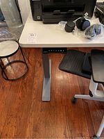 Image of the desk with white tabletop and grey legs, showing the control panel on the front side with preset buttons labeled 1, 2, 3, M, and up/down arrows. Some objects on the tabletop including caster wheels and other items are visible.