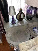 Two grey wooden end tables each with a curved side storage drawer, a lower tier shelf, and decorative glass-look knobs. Both have a distressed finish and carved legs. The right one shown with a purple lamp and vase on top.
