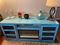 Front view of turquoise wooden entertainment center with glass-paneled doors and electric fireplace insert, decorated with lamp, glass jar, and other small items on top.