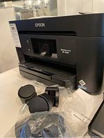 Front-side view of a black Epson Workforce Pro WF-3820 printer with four caster wheels in front wrapped in plastic and loose, placed on a white surface with chandelier reflection in background.