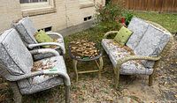 Full view of the wicker loveseat, two chairs, and glass top table with cushions and green throw pillows outside, showing wear and dirt.