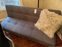 Futon couch in grey fabric with tufted seats and backrest, accompanied by a shaggy grey textured pillow.