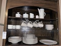 Wide view of all dinnerware and serveware pieces arranged on wooden shelves including plates of various sizes, cups, mugs, a creamer, sugar bowl, and coffee pot all white with garland embossing.