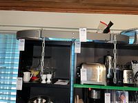 Metal hanging pot rack with attached chains, glass cake plates/stands, and small kitchenware displayed on a black shelf in a home environment.