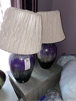 Photo of two ceramic table lamps with purple, grey, and silver bases and grey pleated lampshades on a table.