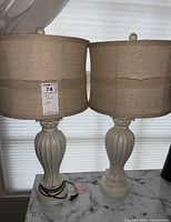 Two similar lamps standing side by side on a marble surface in front of a window with blinds. Each lamp has a beige linen drum shade and a white cream distressed base with carved vertical grooves.