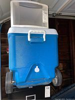 Blue Rubbermaid Dura Chill cooler viewed from front with large wheels, white handle, and white drain spout.