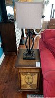 Wooden side table with palm and leaf motif on door panel, metal lamp with grey shade on top, next to red couch