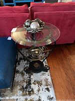 Side table next to sofas showing full height and decorative metal base with spherical design elements, glass top clear with brass-colored accent; decorative bowl with four spheres on top.