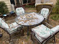 Five metal Chairs with floral cushions surrounding a round glass-top table on metal base, outdoor setting with fallen leaves.
