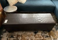 Photo showing full length and top of dark brown storage bench with stitched detail and label attached.