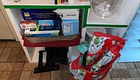 Photo of two black TV wall mounts, two boxes labeled TV mounts, two glass beverage dispensers, reusable shopping bags with red accents, DVDs, and various household items arranged on shelves and on the floor.