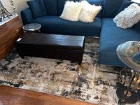 Large 8x10 area rug underneath dark blue sectional sofa and brown leather bench, showing abstract brown, gold, black and beige tones on white base.