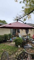 Umbrella fully extended outside near patio furniture and garden plants, dark red canopy with some visible wear and fading.