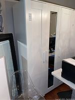 White wooden armoire with a mirror door and two vertical handles, shown from an angle with nearby furniture visible.