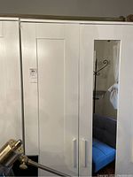 Photo showing white wooden armoire with vertical panel doors, one door with full-length mirror and white handles.