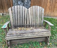 Front view of weathered wooden double glider bench with vertical slats and armrests, placed outdoors on grass, showing wear and leaf debris.