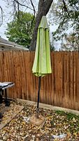Outdoor umbrella with light green canopy attached to a metal pole and square weighted base, situated in backyard near wooden fence with fallen leaves on ground.