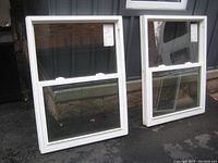 Two white framed vinyl sliding windows standing outdoors, one appears fully intact and the other missing window hinges and some hardware parts.