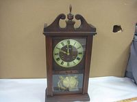 Full front view of the wooden windup wall clock with glass panels showing dial and pendulum.