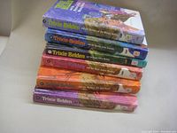 Photo showing the spines of six Trixie Belden hardcover books stacked horizontally. Each spine shows unique title and series branding, with some wear on the edges.