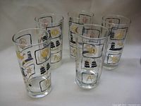 Set of five clear high ball glasses with nautical designs including anchors, lighthouses, ship wheels and seashells in black and gold and tan colors.