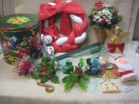 Photo showing assorted Christmas decorations arranged together including a fabric wreath, tin can, snowman figurine, basket with poinsettias, angel figurine, and various greenery and ornaments.