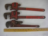 Three red preowned pipe wrenches laid out with ruler for scale, one Benchmark measuring 14 inches and two Super Ego wrenches measuring 18 inches each.