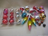 Full view of the 27 vintage glass Christmas ornaments arranged on a beige surface, showing variety of shapes and colors