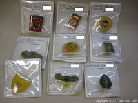 Top view of nine vintage military style pins in their original sealed clear plastic packages with labeling cards showing unit names and some measurements.