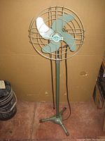 Front view of the vintage Sea Breeze standing fan showing green blades inside a protective wire cage and supported on a green metal stand with three legs.