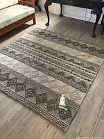 Square area rug laid out on tiled floor showing geometric diamond and stripe patterns in neutral tones.