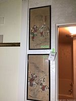 Photo showing two framed Asian fabric art prints hanging on a wall, one above the other, with measuring tape and lot tag visible.