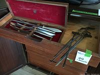 Wooden case with red felt interior open displaying stainless steel BBQ knives and forks with wooden handles next to metal BBQ skewers.