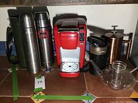 Lot items including red Keurig coffee maker, thermoses, coffee grinder, glass tea kettle, and bronze coffee press laid out on surface.