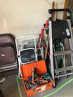 Photo showing multiple folding ladders, an orange toolbox with assorted hand tools, and gardening/camping tools leaning against wall.
