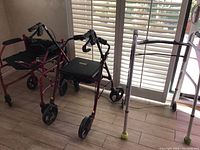 Photo showing three mobility assistance devices: a burgundy 4-wheel rollator walker with seat, a silver 4-legged walker with yellow leg tips, and a lightweight manual wheelchair with black fabric seating and armrests.
