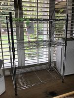 Metal storage rack with three wire shelves placed near window with blinds in background.