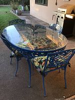 Full view of the oval glass-top metal patio table with four blue metal chairs around it, showing detailed ornate design on chair backs and colorful cushions.