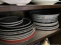 Shelving with stacks of plates featuring white bases with red and blue rims, and dark ceramic plates with a brown abstract design.