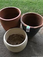 Three large pots planters shown outdoors on concrete and grass. Pots vary in color and style with some soil residue inside the smallest pot.
