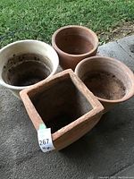 Four heavy garden pots and planters arranged outside on pavement near grass. Includes one large square terra cotta planter and three round pots.