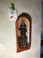 Full view of the painted wooden plaque displaying the monk figure with a red and yellow ornamental border, hanging on a wall.
