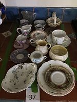 Photo of multiple teacups, saucers, cream pitchers, and decorative basket arranged on table.